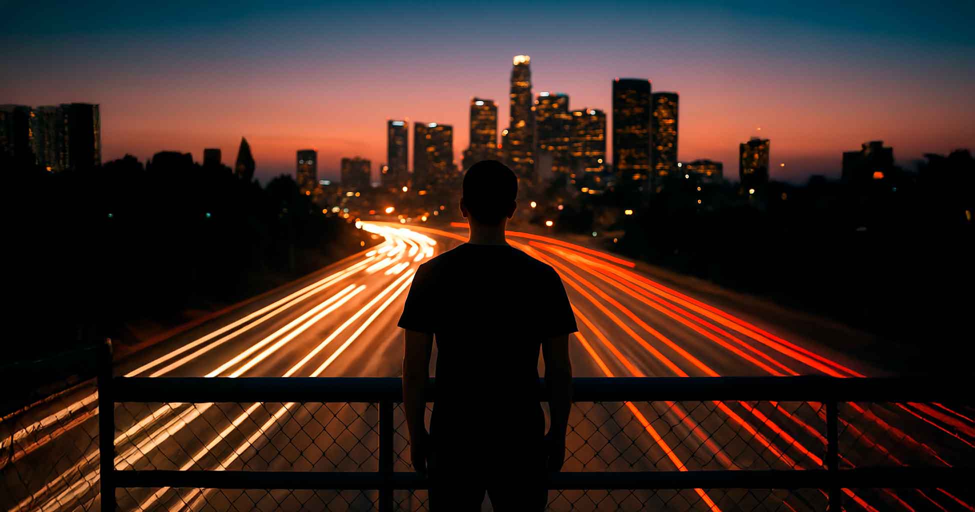 Still Training – Everyday Practice in a Moving World A lone man stands on an overpass at twilight, watching traffic light trails flow beneath him, with a city skyline glowing in the background.